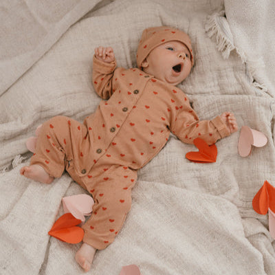 Een baby in een koraalkleurig boxpakje met rode hartjes en een bijpassend babymutsje