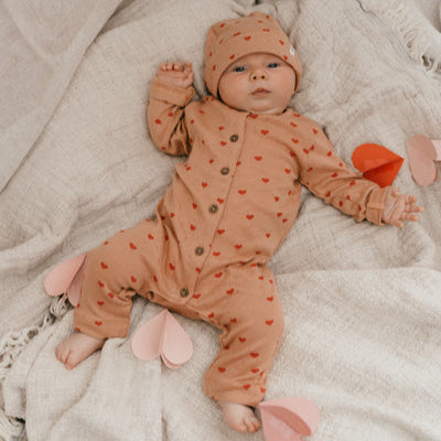 Een baby in een koraalkleurig boxpakje met rode hartjes en een bijpassend mutsje