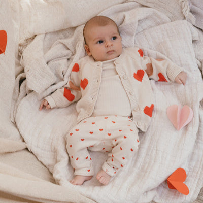 Een baby in een gebroken wit babyvestje met hartjes een ivory kleurige longsleeve en een gebroken wit broekje met rode hartjes