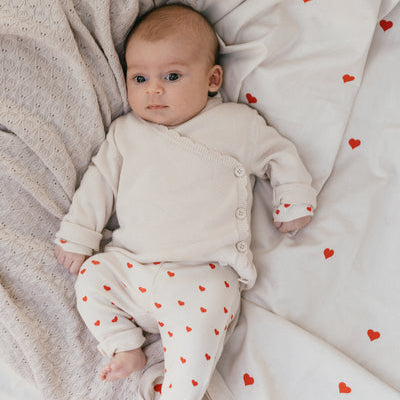 Een baby in een gebroken wit overslagshirtje met een gebroken wit broekje met rode hartjes