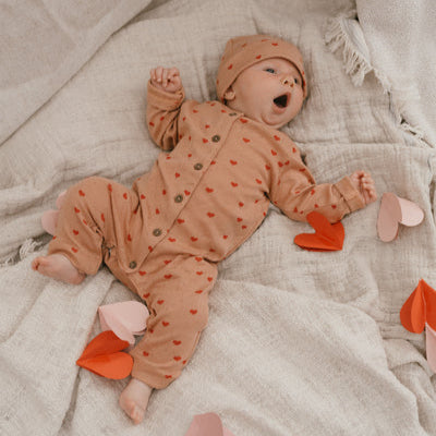 Een babytje in een koraalkleurig boxpakje met rode hartjes en een bijpassend mutsje
