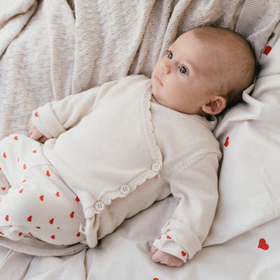 Een baby in een gebroken wit overslagshirtje met een gebroken wit broekje met rode hartjes