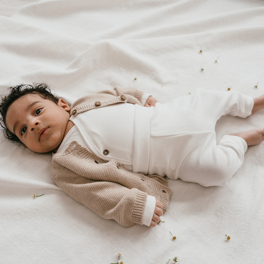 Een baby in een beige vestje, witte overslagromper en een wit broekje