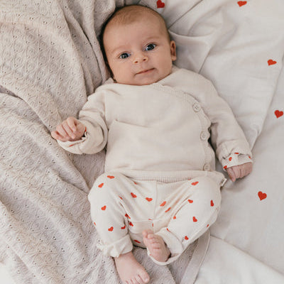 Een baby in een gebroken wit overslagshirtje met een gebroken wit broekje met rode hartjes