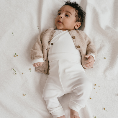 Een baby in een beige vestje, witte overslag romper en wit broekje