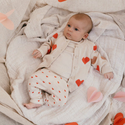 Een baby in een gebroken wit babyvestje met hartjes een ivory kleurige longsleeve en een gebroken wit broekje met rode hartjes