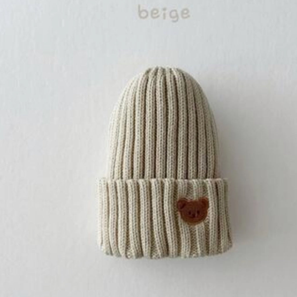Een beige muts met een beertje er op geborduurd.