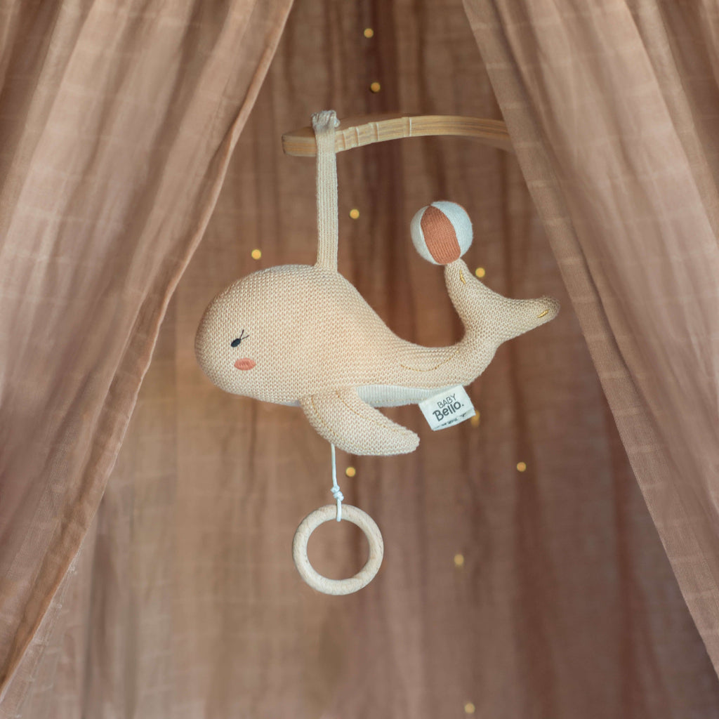 Een beige kleurige muziekmobiel in de vorm van een walvis hangend in een ledikantje