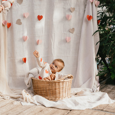 Een kindje in een rietenmandje met een ivory kleurig babyvestje met hartjes en een koraalkleurig t-shirt