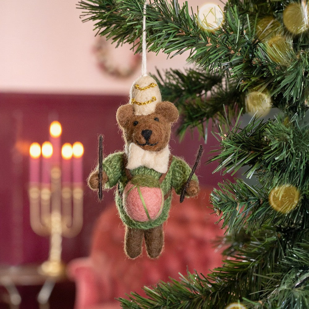 Een kerstboomhanger een bruin beertje met beige muts en strik in een groene trui en roze trommel. Gemaakt van wolvilt.