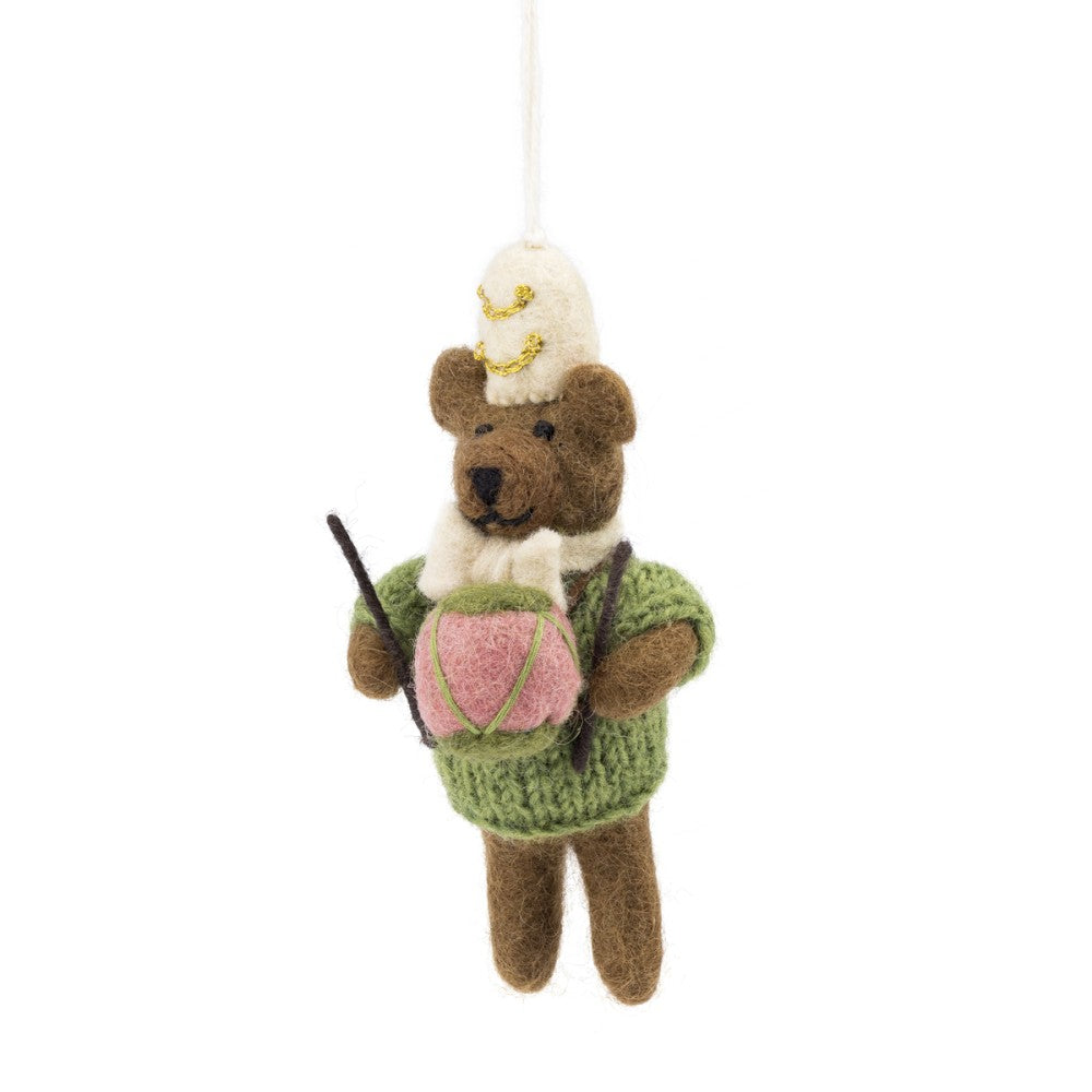Een kerstboomhanger een bruin beertje met beige muts en strik in een groene trui en roze trommel. Gemaakt van wolvilt.