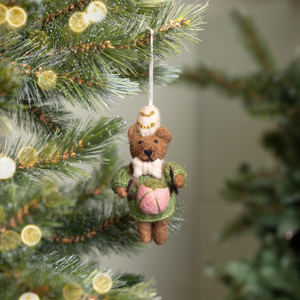 Een kerstboomhanger een bruin beertje met beige muts en strik in een groene trui en roze trommel. Gemaakt van wolvilt.