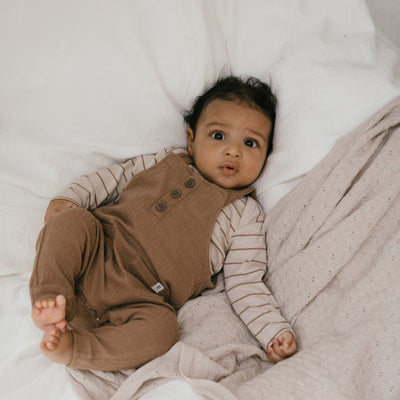 Een baby in een bruine tuinbroek met een beige longsleeve met streepjes